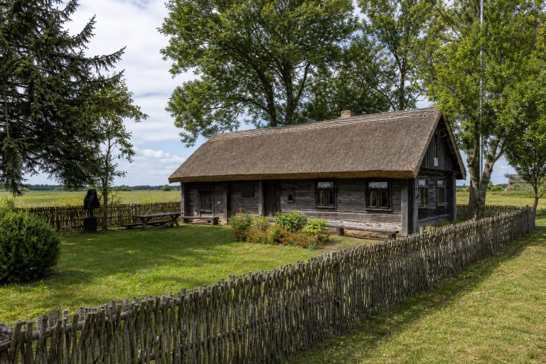 Tamsių rąstų Juozo Zikaro memorialinė sodyba Paliukuose su nendriniu stogu, pinučių tvora, gėlėmis ir brandžiais medžiais