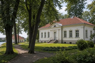 Light-coloured late Classicist service wing with a red roof among trees in Jurbarkas Manor Park