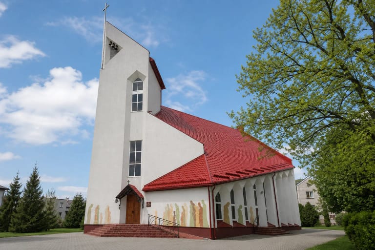 Balta Jurbarko Kristijono Donelaičio evangelikų liuteronų bažnyčia su aukštu bokštu, raudonu stogu ir spalvotais žmonių siluetais fasade