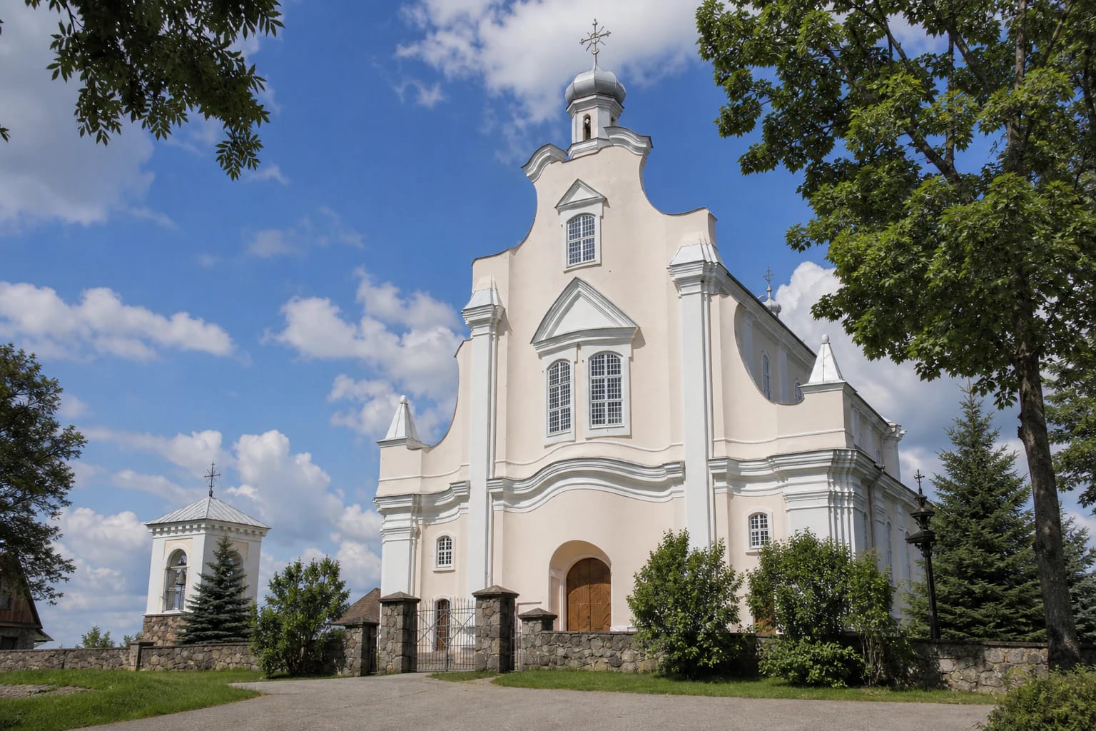 Pale curving façade of Jūžintai Church of St Michael the Archangel with its fieldstone wall and separate white bell tower in summer