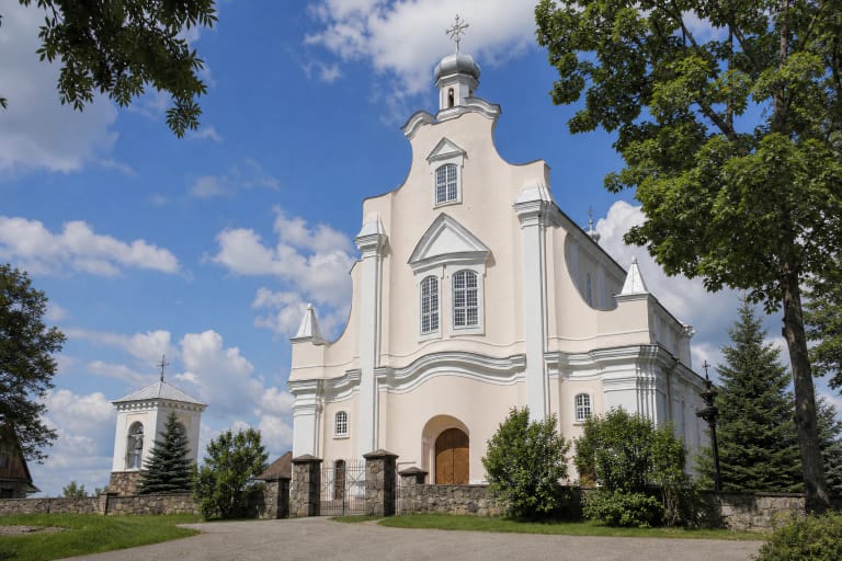 Lenktų linijų Jūžintų Šv. arkangelo Mykolo bažnyčios fasadas, akmenų tvora ir atskira balta varpinė vasarą
