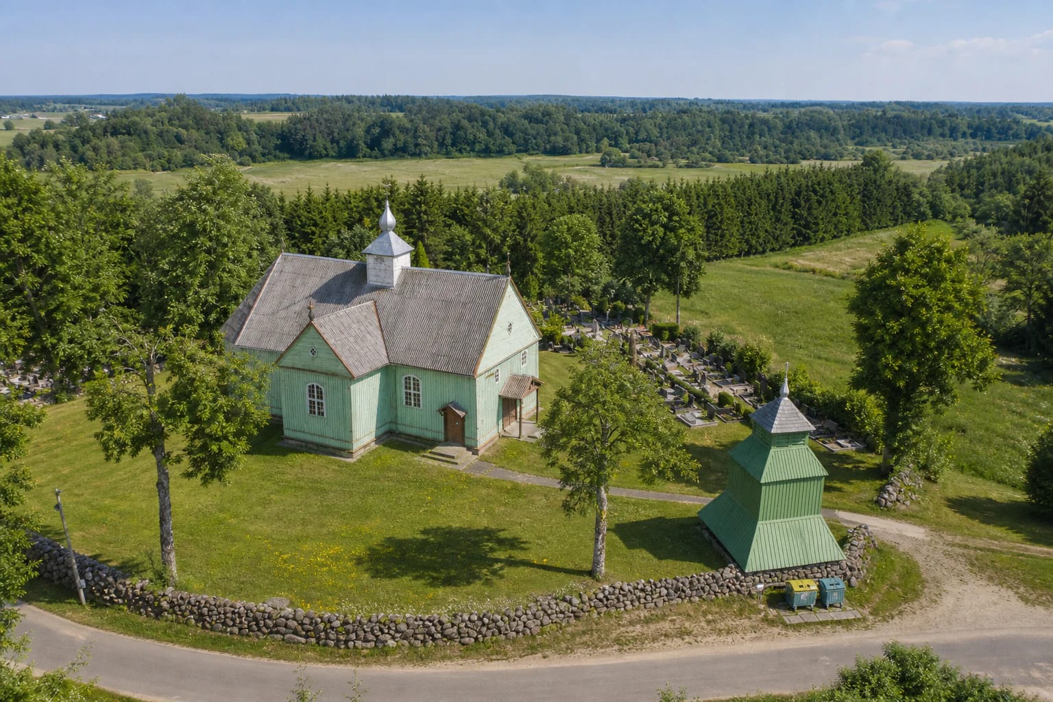 Pale-green timber Church of St Lawrence at Kalnalis, its separate three-tier bell tower, and fieldstone wall above the Salantas valley