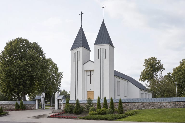 Balta dvibokštė Kaltinėnų Šv. Jono Krikštytojo bažnyčia už akmenų tvoros