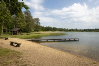 Sandy recreation area on Lake Kapstatas with a timber pontoon bridge, shelter, and low wooded shore