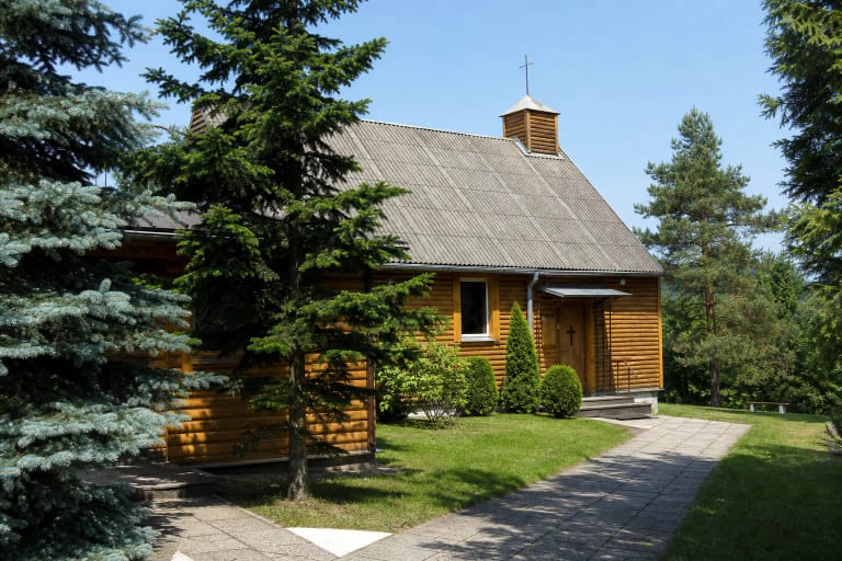 Kukli medinė Karmėlavos Šv. Onos bažnyčia su rusva lentų apdaila, plačiu pilku stogu ir mažu bokšteliu tarp eglių