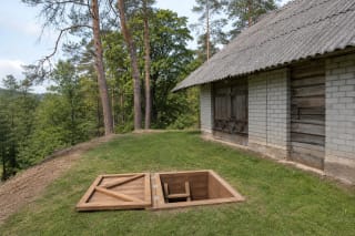 открытый деревянный люк бункера партизанской штаб-квартиры в Касчюнай на лугу у старой хозяйственной постройки и лесистого обрыва Меркиса