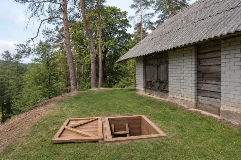 Atvertas medinis Kasčiūnų partizanų vadavietės bunkerio liukas pievoje prie seno ūkinio pastato ir miškingo Merkio skardžio