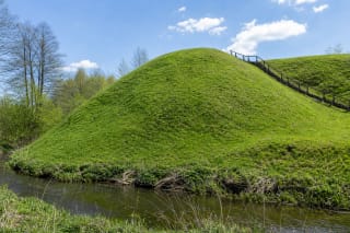 Steep grass-covered bank of Kaukai Hillfort beside the Peršėkė with timber steps under a blue spring sky