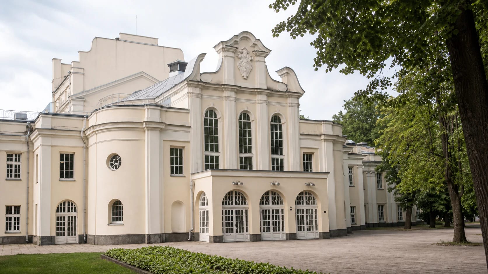 Cream-coloured Neo-Baroque facade of Kaunas State Musical Theatre in the City Garden with three arched windows, curved pediment, and trees