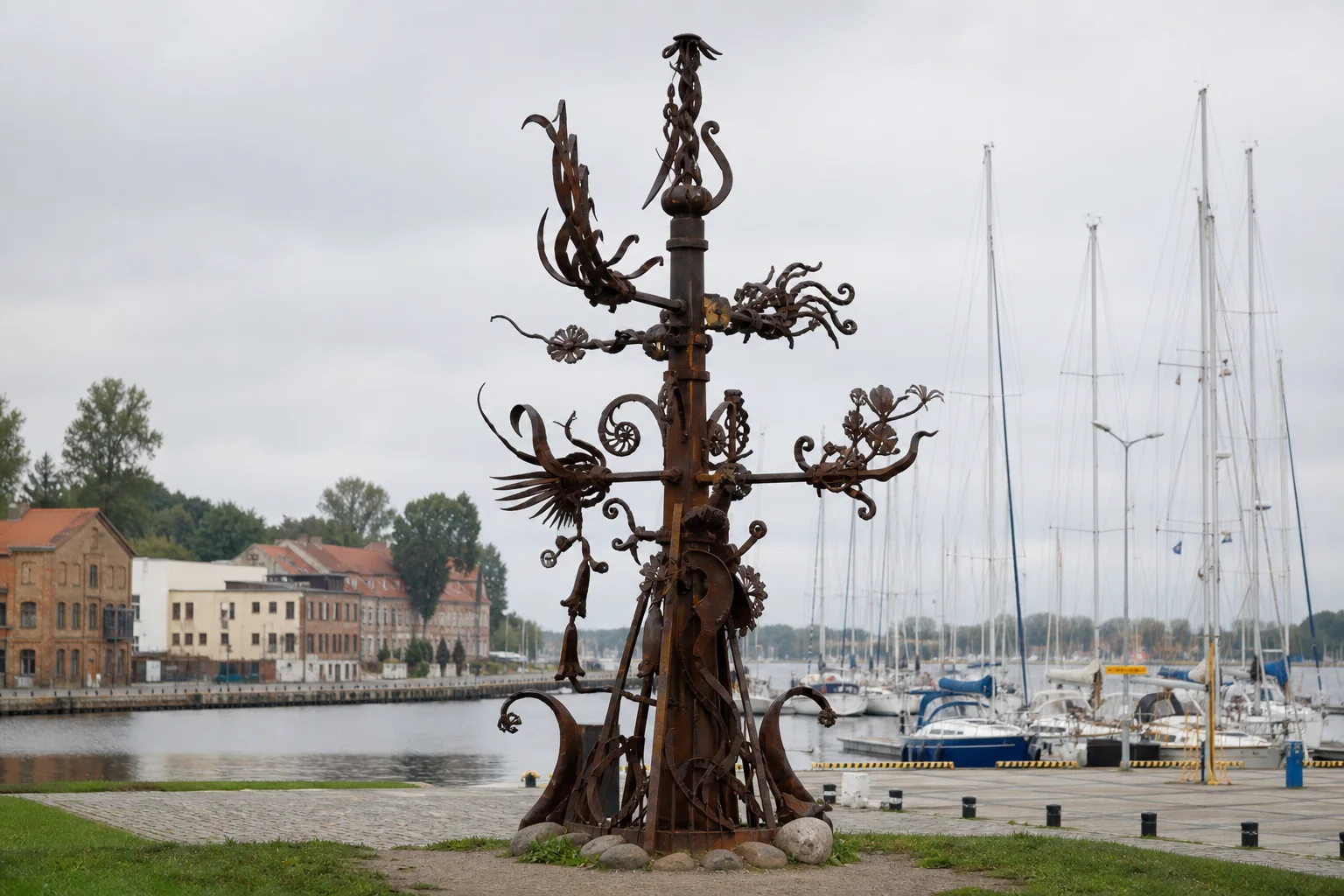 Tall rust-coloured Four Winds forged-metal composition with curling details against Klaipėda Castle Harbour