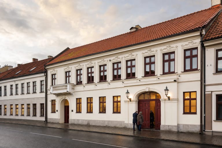 Klaipėdos koncertų salės istorinis šviesus fasadas su tamsiai raudonais langais ir čerpiniu stogu po lietaus