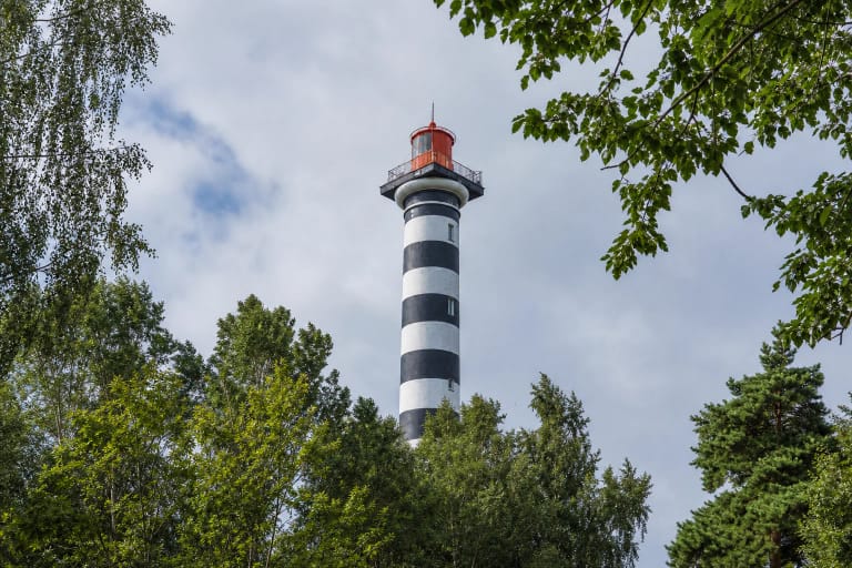 Juodomis ir baltomis horizontaliomis juostomis dažytas Klaipėdos švyturys su raudona žibinto patalpa virš Melnragės medžių