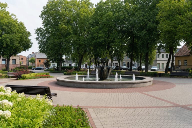 Apskritas veikiantis fontanas Knypavos Rinkos aikštėje, apsuptas šviesaus grindinio, gėlynų, suolų ir brandžių medžių