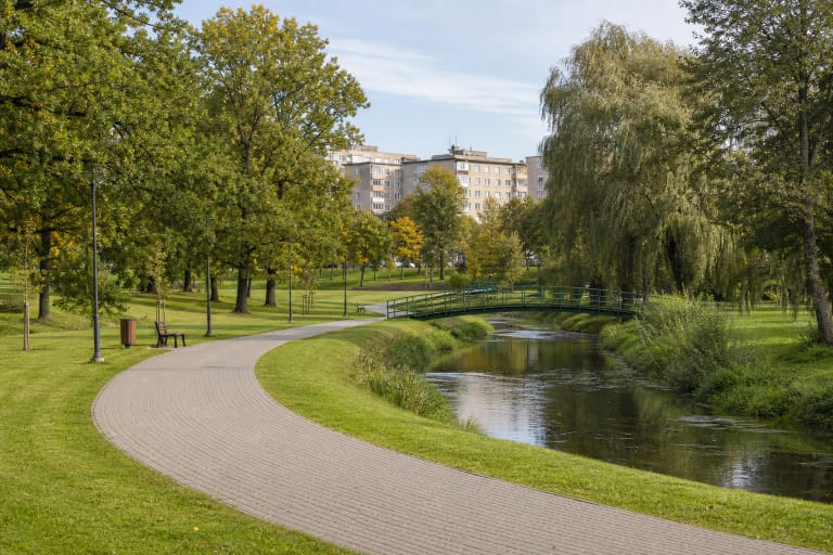 Krašuonos upelis, vingiuotas grįstas takas, žalias metalinis tiltelis ir ąžuolų veja Krašuonos parke Utenoje