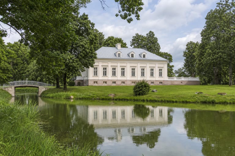 Krikštėnų dvaro šviesūs rūmai, balustrados terasa, žalias parkas, metalinis tiltelis ir pastatą atspindintis tvenkinys