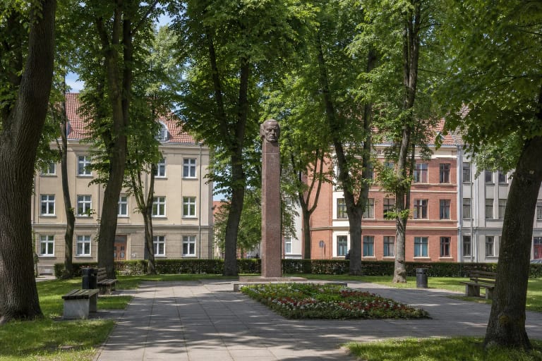 Kristijono Donelaičio aikštės raudono granito paminklas tarp brandžių liepų ir istorinių Klaipėdos Naujamiesčio namų