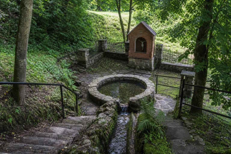 Stone basin and channel of Krokulė Spring in a green hollow, with a red-brick chapel and stone steps nearby
