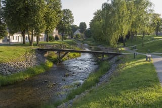 Timber footbridge over the narrow Kupa in a green Kupiškis valley park with stone-reinforced banks