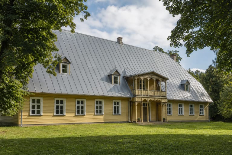 Светло-жёлтая деревянная усадьба Куршенай с серой крышей и резным балконом в зелёном парке