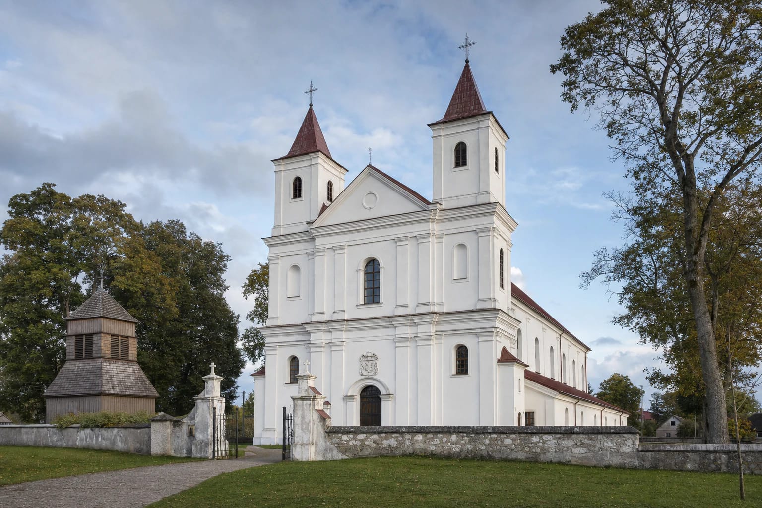 White twin-towered Kurtuvėnai Church of St James with red roofs, a timber belfry, and a masonry churchyard wall in a Žemaitija town