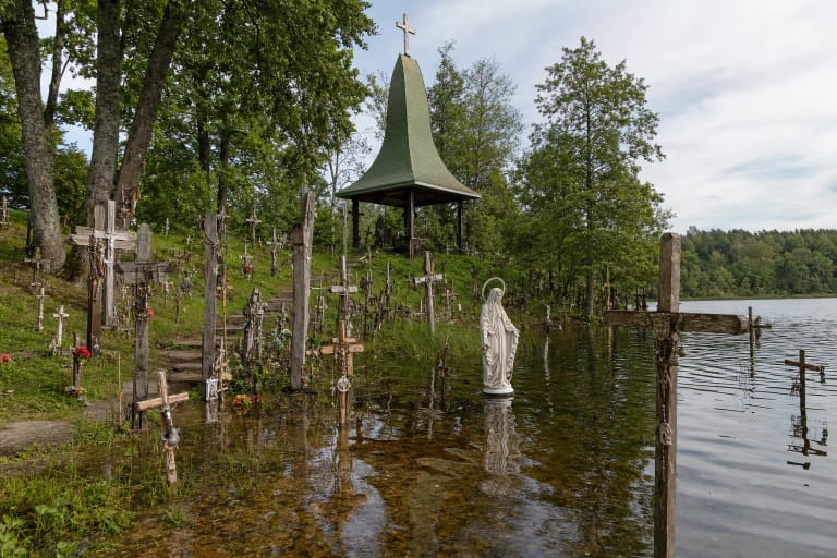Kvintiškių šventvietės kryžiai ir Marijos statula Ilgio ežero vandenyje, o virš pakrantės kyla koplyčia aukštu žaliu stogu