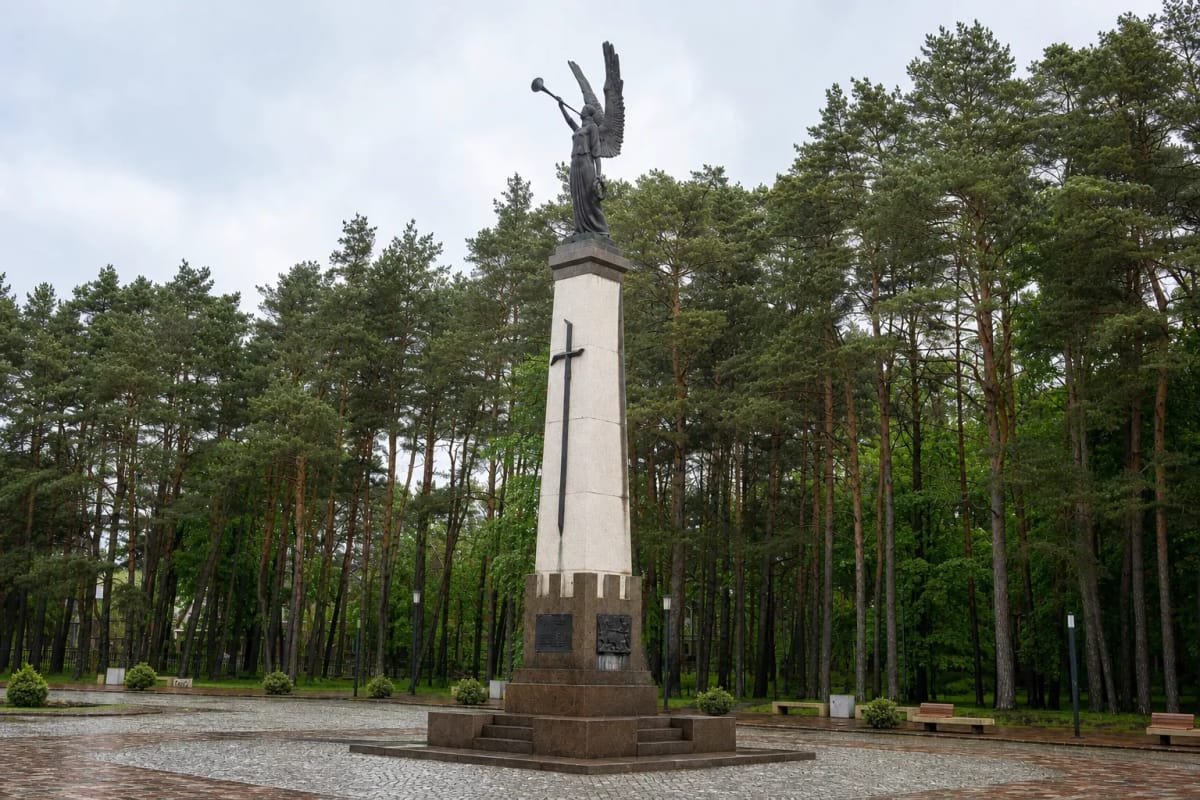 Бронзовый трубящий «Ангел свободы» на высоком светлом обелиске среди сосен на широкой площади Алитуса