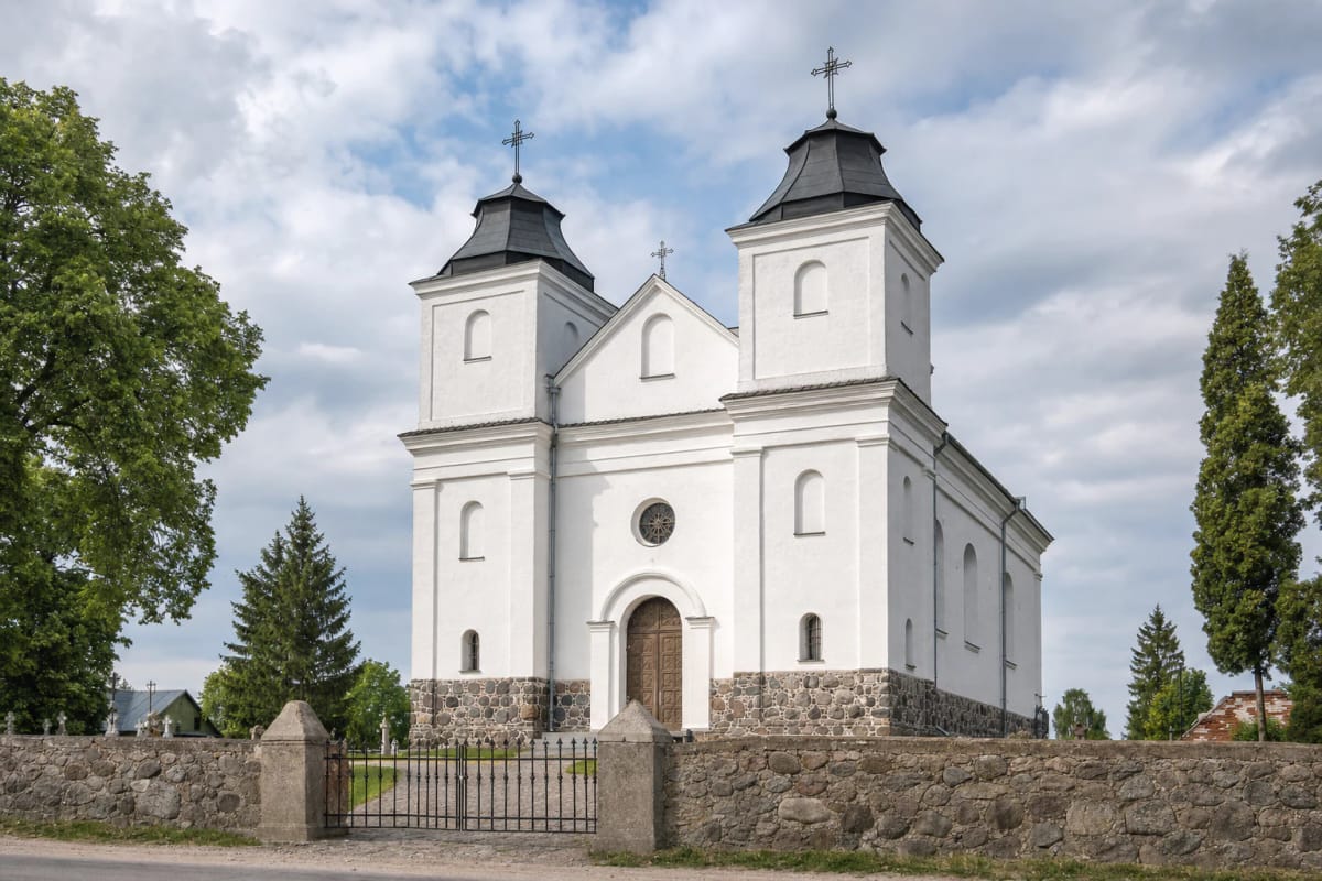 White twin-tower Church of the Discovery of the Holy Cross in Laukuva behind a fieldstone wall and iron gate