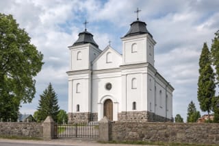 Balta dvibokštė Laukuvos Šv. Kryžiaus Atradimo bažnyčia su žemais tamsiais bokštų stogais už lauko akmenų tvoros ir geležinių vartų