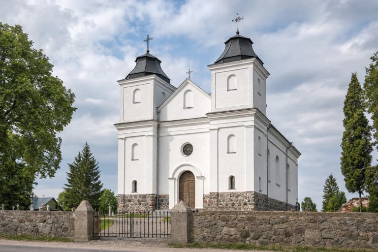 Balta dvibokštė Laukuvos Šv. Kryžiaus Atradimo bažnyčia su žemais tamsiais bokštų stogais už lauko akmenų tvoros ir geležinių vartų