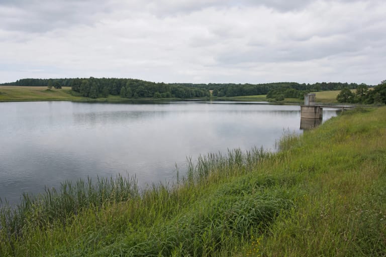 Calm water of Laukžemiai Reservoir with a green shore, woodland, fields, and a concrete water-control structure