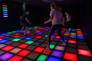 Three players moving across red, green, and blue illuminated sensor tiles in the LAVA FLOOR arena