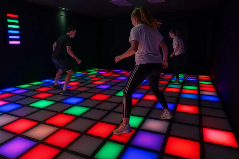 Trys žaidėjai juda per raudonai, žaliai ir mėlynai šviečiančias LAVA FLOOR jutiklių grindis