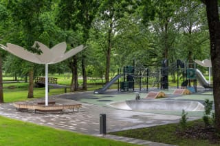 Green and black playground in Lazdijai City Park with climbing nets, a ramp, and white tulip-shaped canopies beneath mature trees