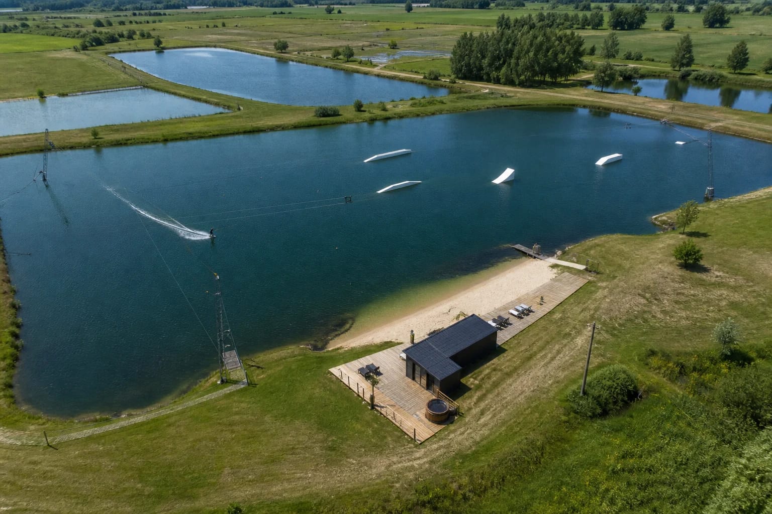 Liberty Beach artificial pond with cable wake course, white obstacles, sandy shore and dark sauna building