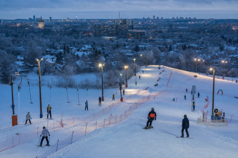 Floodlit urban ski runs at Liepkalnis with surface lifts, a snow park, and the distant Vilnius skyline
