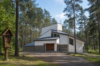 White Church of the Lithuanian Martyrs with angular grey roofs among Berčiūnai pines and a carved wooden Station of the Cross in front