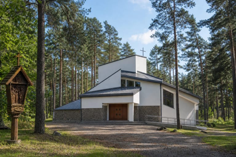White Church of the Lithuanian Martyrs with angular grey roofs among Berčiūnai pines and a carved wooden Station of the Cross in front