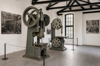 A restored grey-green press, vertical band saw and historic photographs in the white-walled Paul Lindenau shipyard workshop exhibition