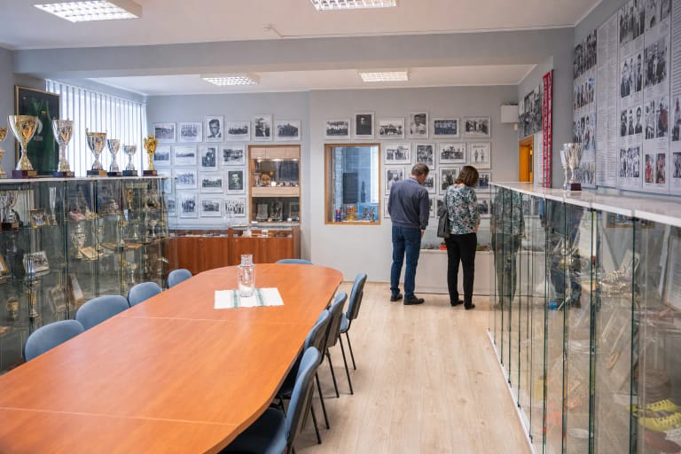Kompaktiška Marijampolės sporto istorijos muziejaus salė su trofėjų vitrinomis, senų sporto nuotraukų sienomis ir centriniu mediniu stalu
