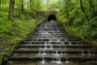 Marvelė stream flowing over broad stone steps beneath the arch of a historic bridge in a green Kaunas valley