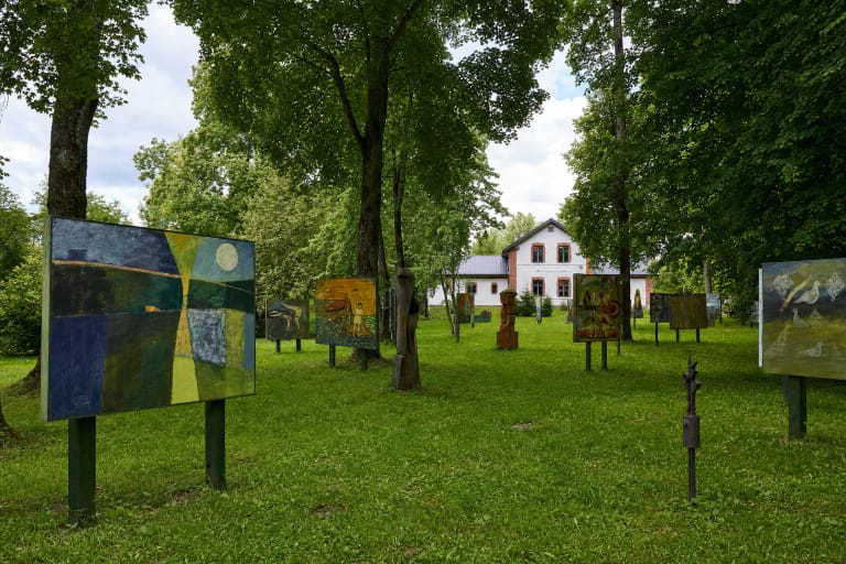 Dideli spalvingi paveikslai ant stovų Mažosios Lietuvos paveikslų sodo pievoje tarp brandžių medžių