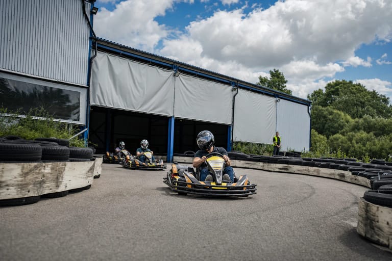 Helmeted drivers in MėmelGP karts emerge from the covered circuit onto outdoor asphalt between tyre barriers