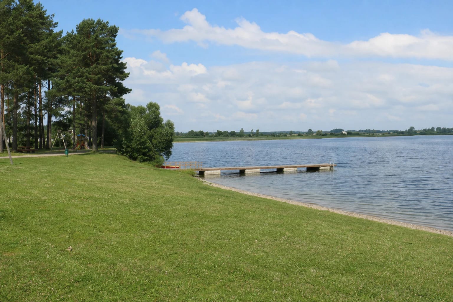 Grassy and sandy Lake Metelys bathing shore with pines, a long low pier, and an open lake horizon