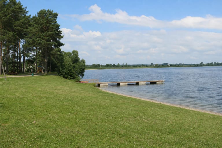 Grassy and sandy Lake Metelys bathing shore with pines, a long low pier, and an open lake horizon