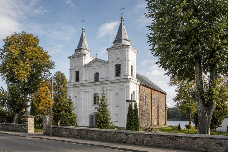Šviesi Metelių bažnyčia su dviem bokštais, centriniu frontonu ir akmenų tvora