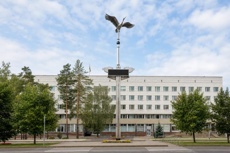 Sidabro spalvos išskleistų sparnų gervė ant aukštos stelos su švieslente priešais Visagino savivaldybės pastatą
