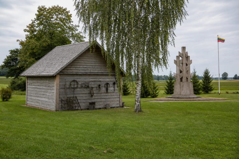 Reconstructed grey timber granary above the Minaičiai partisan bunker and the LLKS declaration memorial in a rural farmstead landscape