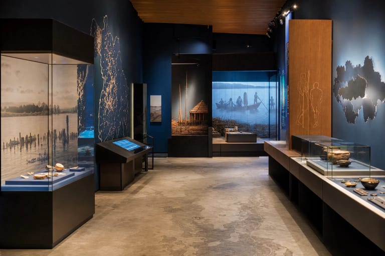 Šiuolaikinė Molėtų krašto muziejaus ekspozicija su archeologijos vitrinomis, vaizdo projekcijomis ir tamsiai mėlynais stendais