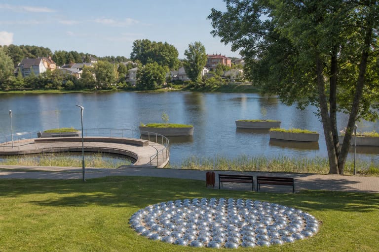 Polished metal hemispheres, green shore, curved timber platform and sculptures in Lake Pastovis at Molėtai Sculpture Park