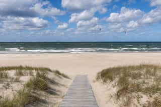 Wooden path through grass-covered Monciškės dunes towards a broad sandy beach, choppy Baltic Sea, and distant kites